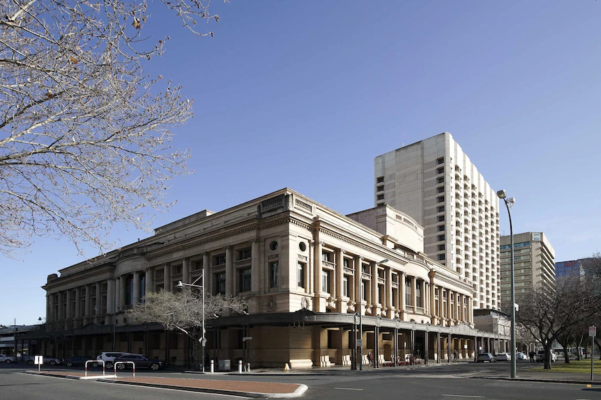 A grand building with columns with a tall building behind and a tree with blossoms in the foreground