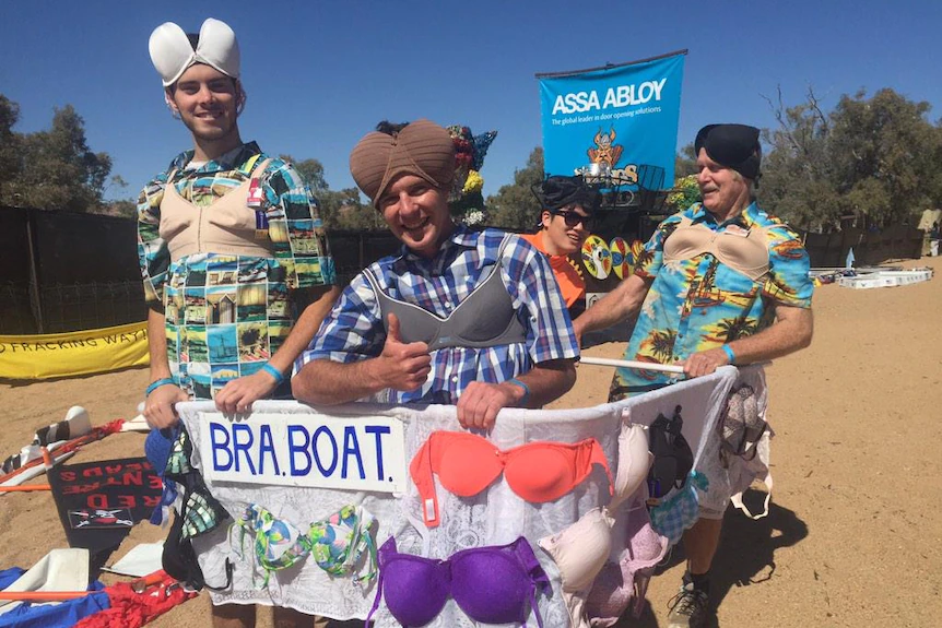 Three men stand in a home-made cardboard boat ready to race down a dry riverbed.